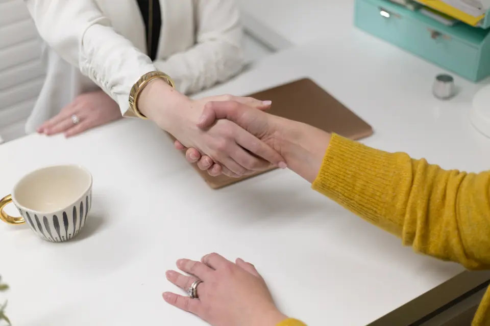 Two people shaking hands across an office desk, a coffee cup beside them.