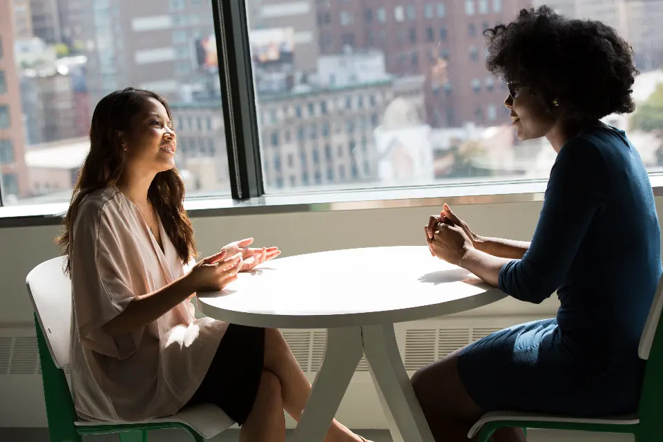 Two women talking across a round table, beside a window with the city behind them.