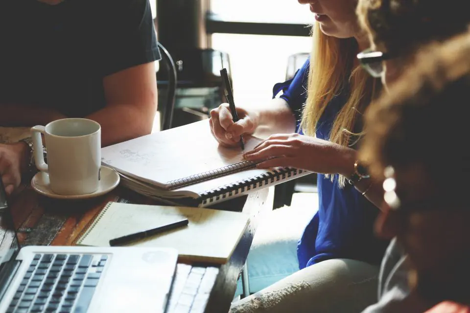 Several people around a work table with open notebooks, coffee cups and a laptop.